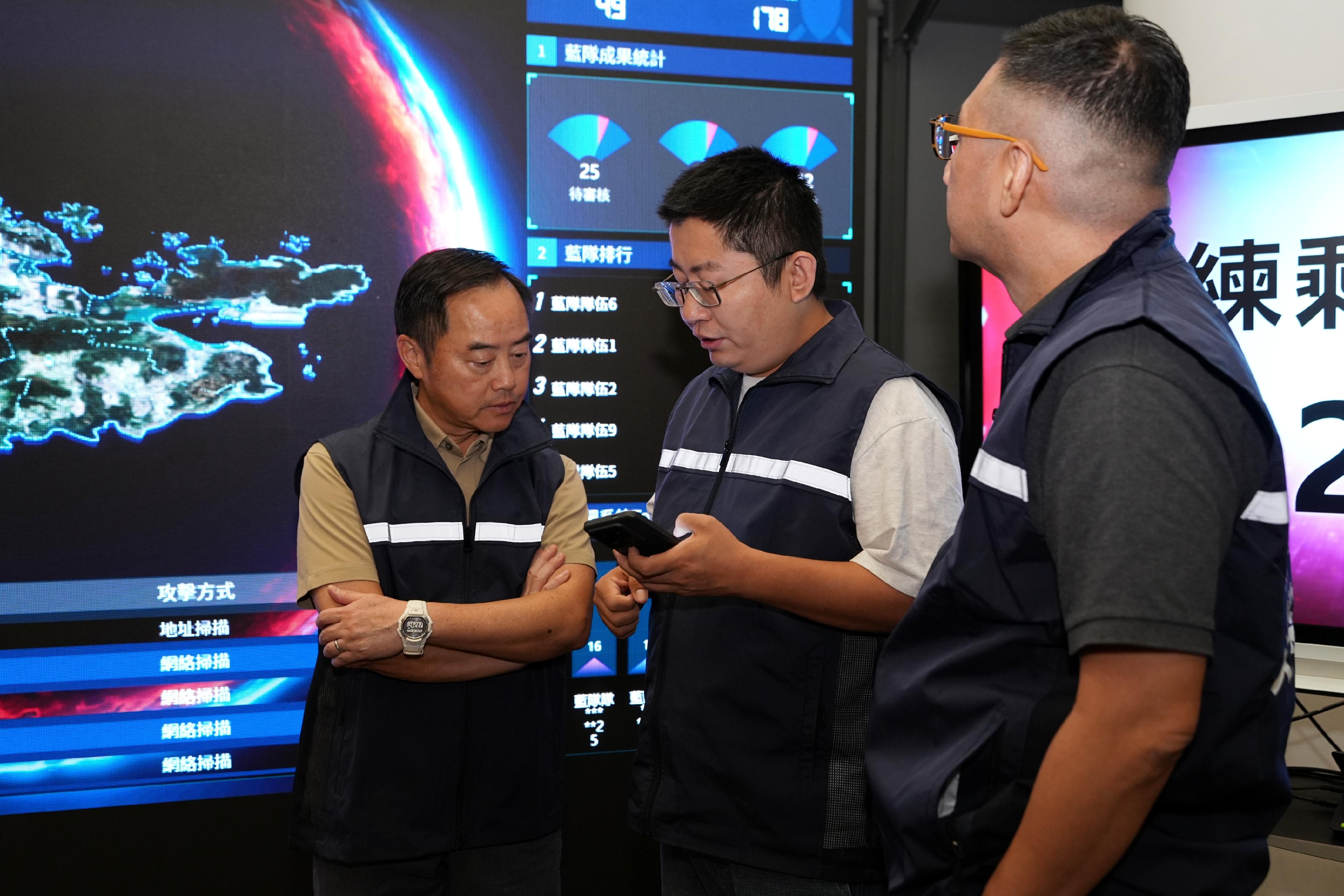 「香港網絡安全攻防演練—以攻築防2025」進行期間，數字政策專員黃志光（左）到場並聽取工作人員介紹活動進度。